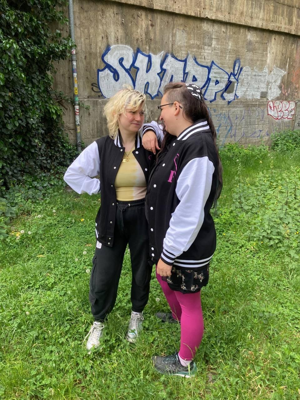 Auf dem Bild sind 2 Menschen zu sehen, die auf einer grünen, saftigen Wiese stehen. Im Hintergrund sind links grüne Pflanzen zu sehen und rechts eine Mauer mit einem Tag. Die Personen sind mittig im Bild. Die eine Person lehnt mit dem Arm auf der Schulter der 2. Person. Die beiden schauen sich in die Augen und lächeln leicht. Beide Personen tragen die gleiche Jacke in schwarz mit weissen Ärmlen und schwarz-weiss gestreiften Bündchen. Die 1. Person trägt ein gelbes T-shirt und schwarze Jogginghosen, weiße Turnschuhe und eine Davidstern Kette. Die Person hat blondierte Haare die vorne kurz sind und hinten bis zur Schulter reichen und zerwuschelt sind. Die 2. Person trägt pinke Leggings und ein schwarzes Kleid mit Blumendruck, schwarze Schuhe und eine Kippah auf dem Kopf. Die Person hat hüftlange Haare und einen Sidecut, eine Brittel und trägt eine Kippah mit Schnurbärten drauf. Beide Person sind askzenasisch-jüdisch und werden osteuropäisch gelesen.